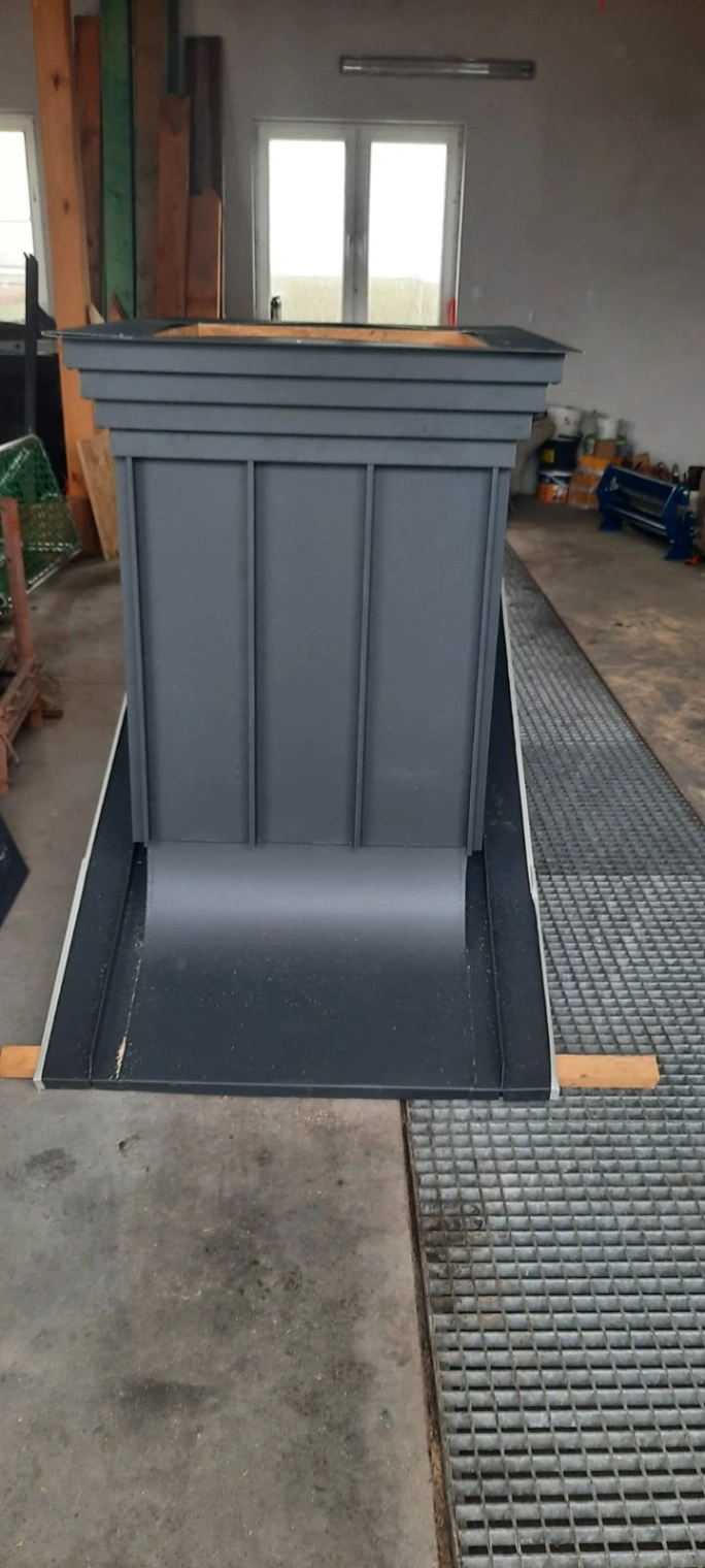 Obróbka komina z czarnej blachy o trapezowej formie ustawiona w warsztacie